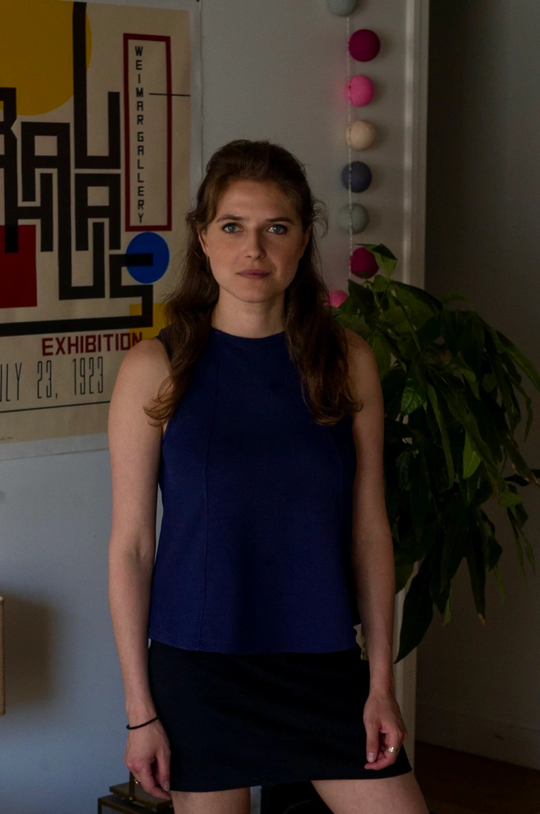 Portrait de Paloma Moritz, débardeur bleu marine, debout devant une affiche Bauhaus et une plante verte